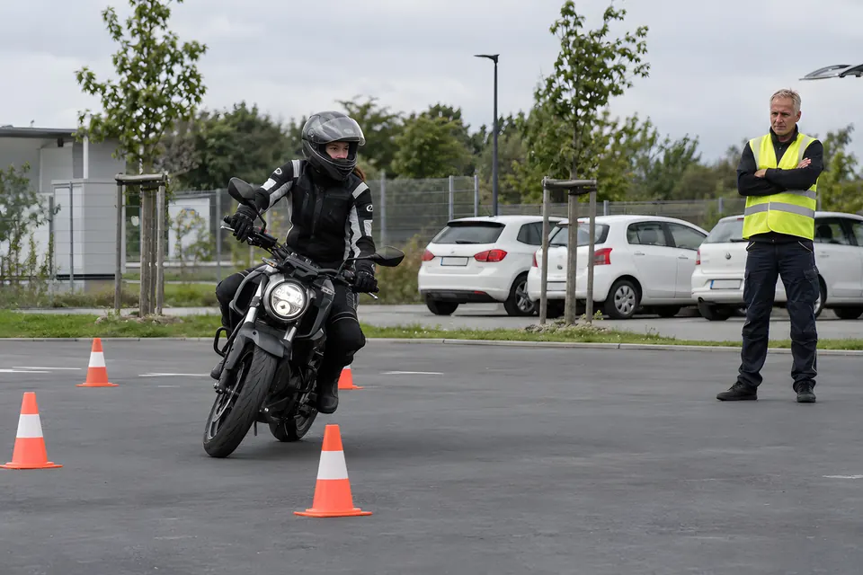 Fahrschüler in Schutzkleidung auf einem Motorrad in einem Übungsparcours mit Pylonen, daneben ein Fahrlehrer in Warnweste