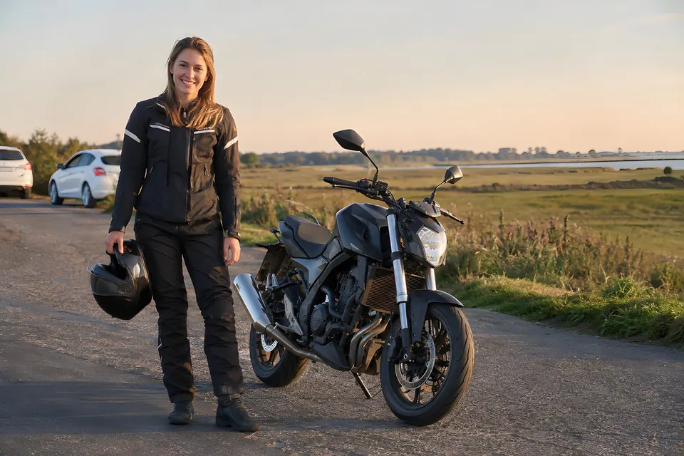 Motorradfahrerin mit Helm in der Hand neben einem Naked Bike im Abendlicht